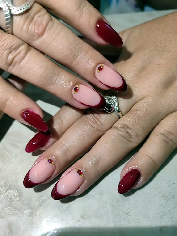 Uñas almendradas con francesa burdeos brillante y pequeños cristales rojos con detalle dorado
