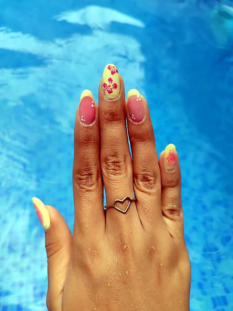 Mano con uñas amarillo pastel y rosa con hibiscos pintados, sobre agua azul